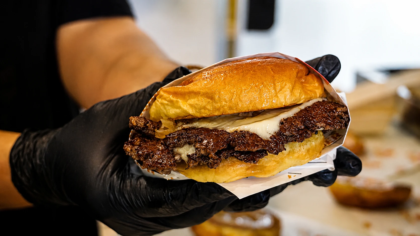 Hand holding a freshly made crispy burger with melted cheese and toasted brioche bun in a commercial kitchen setting.