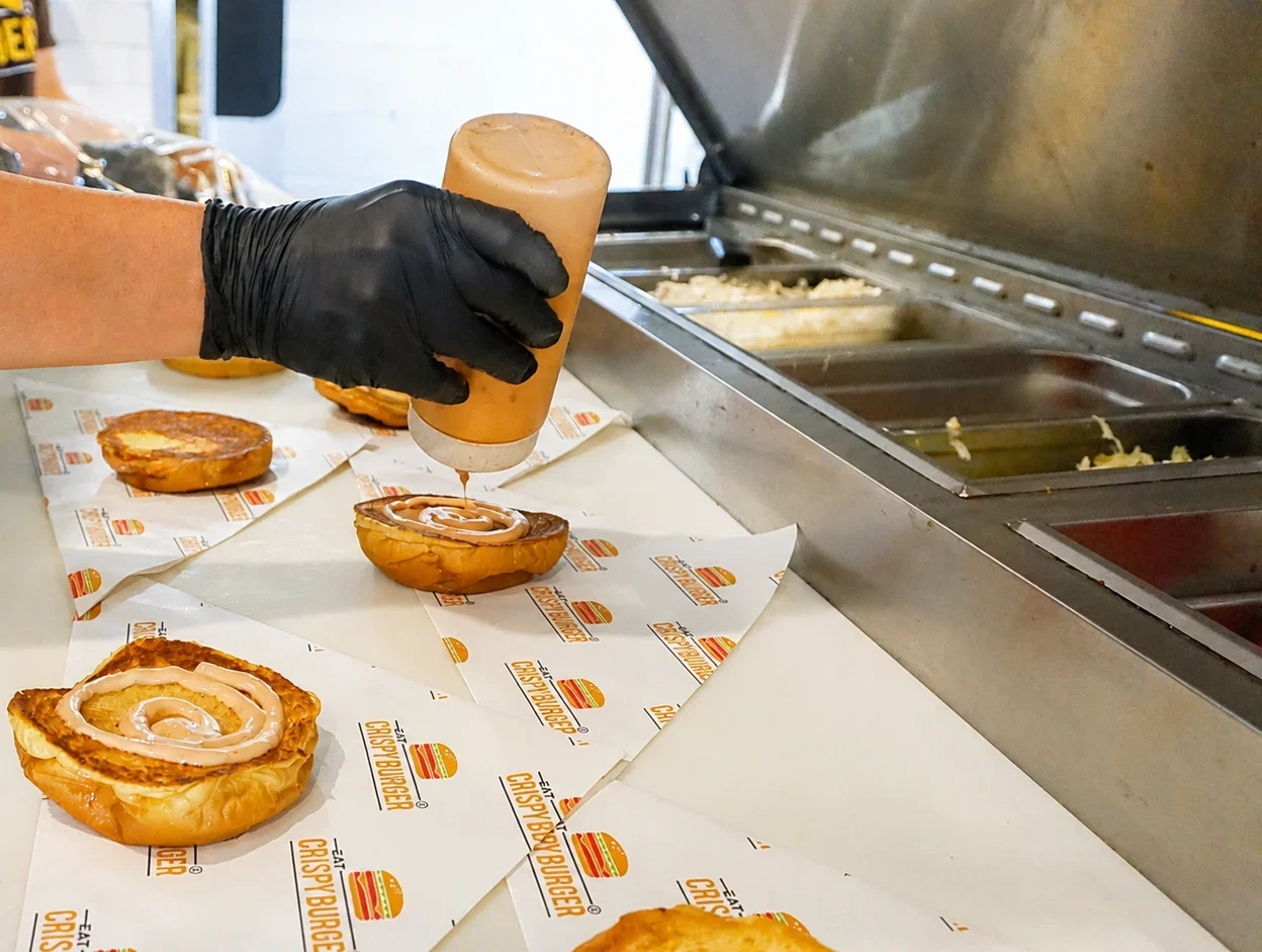 Chef wearing black gloves applying sauce to a toasted burger bun at a restaurant prep station.