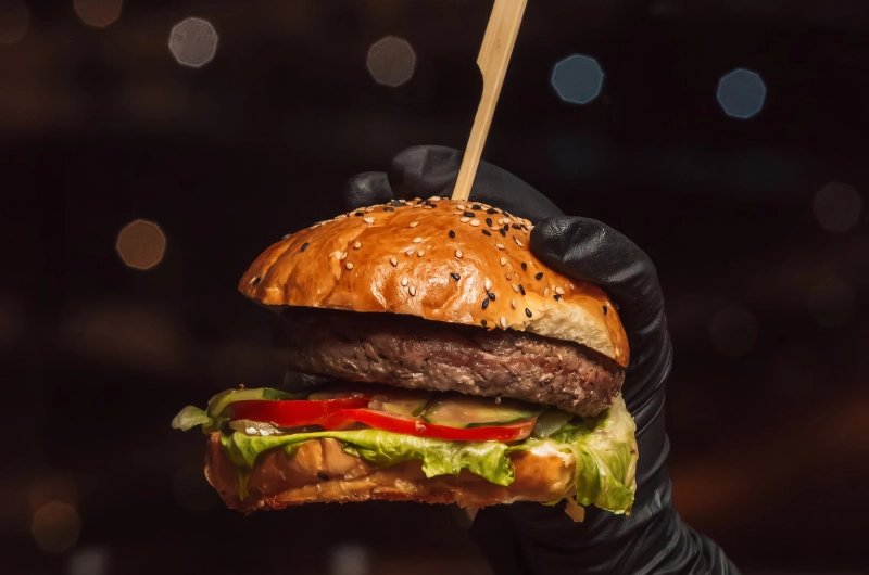 A person in black gloves holds a hamburger, showcasing the meal's layers and toppings.